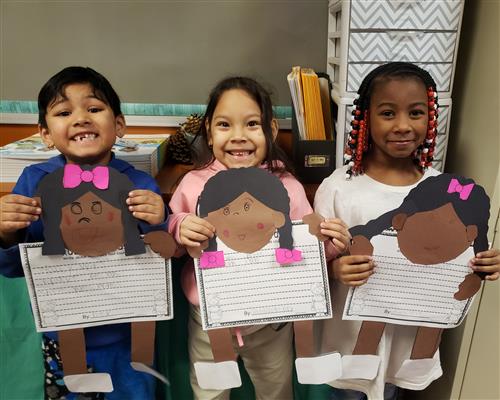 Three smiling students, one boy, two girls, holding up construction paper cutouts of Ruby Bridges.