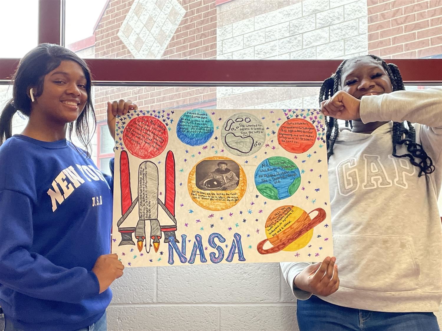 Two female students holding a poster board featuring information about Mae Jemison.
