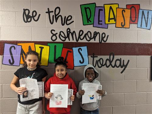 Photo shows three fourth-grade students holding up  written reports they created of famous Black artists or musicians.