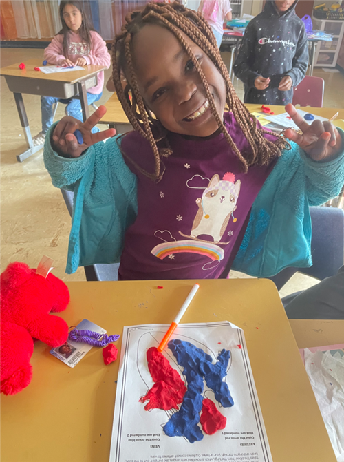 Second grade female student sitting at a desk smiling in front of an art project recreating the heart with colored paper.