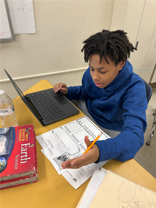 Student sitting at desk, using laptop and paper worksheet to research a famous Black scientist.