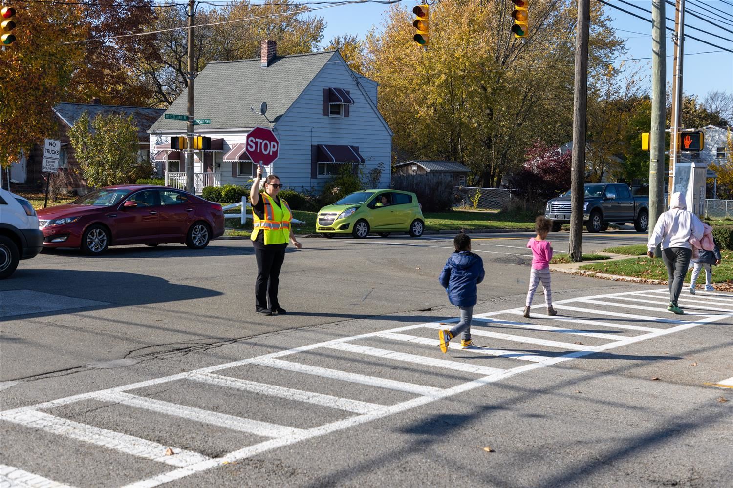 Photo of a crossing guard standing in the middle of a street intersection, holding a stop sign, while 3 children walk across.